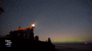 A lighthouse is illuminated and emanates a small wedge of light. The night sky is filled with stars and the Aurora Borealis is visible on the horizon