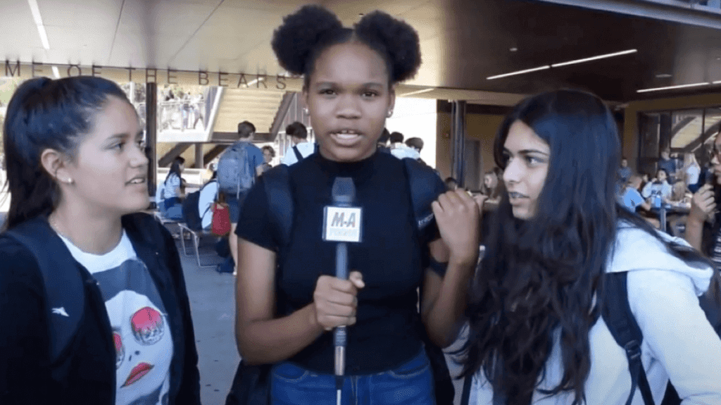 Two teenaged girls talk to a third one who is holding a microphone.