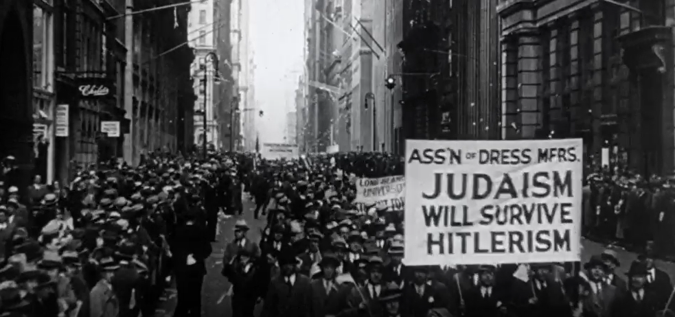 Black and white footage of a crowd of people marching down a city sreet. A group is holding a large sign reading "Ass'n of Dress MFRS. Judaism Will Survive Hitlerism"