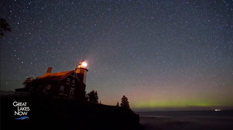 A lighthouse is illuminated and emanates a small wedge of light. The night sky is filled with stars and the Aurora Borealis is visible on the horizon