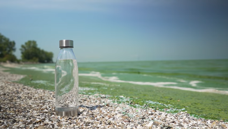 A clear reusable water sits on a pebble beach