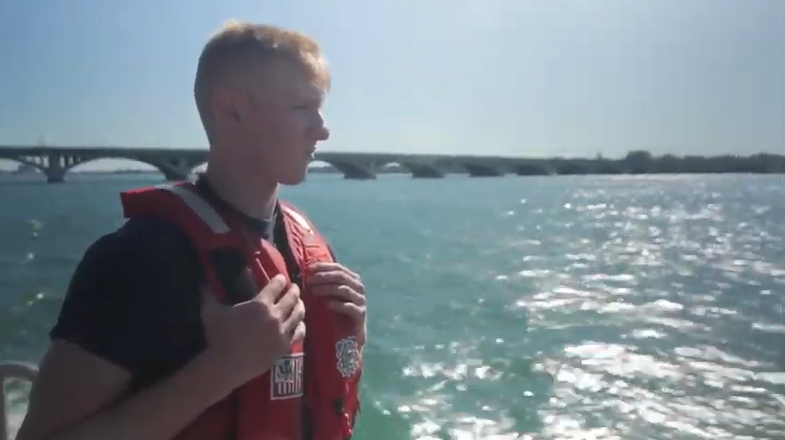 A white man with short blond hair wearing a life vest looks over a lake from a boat