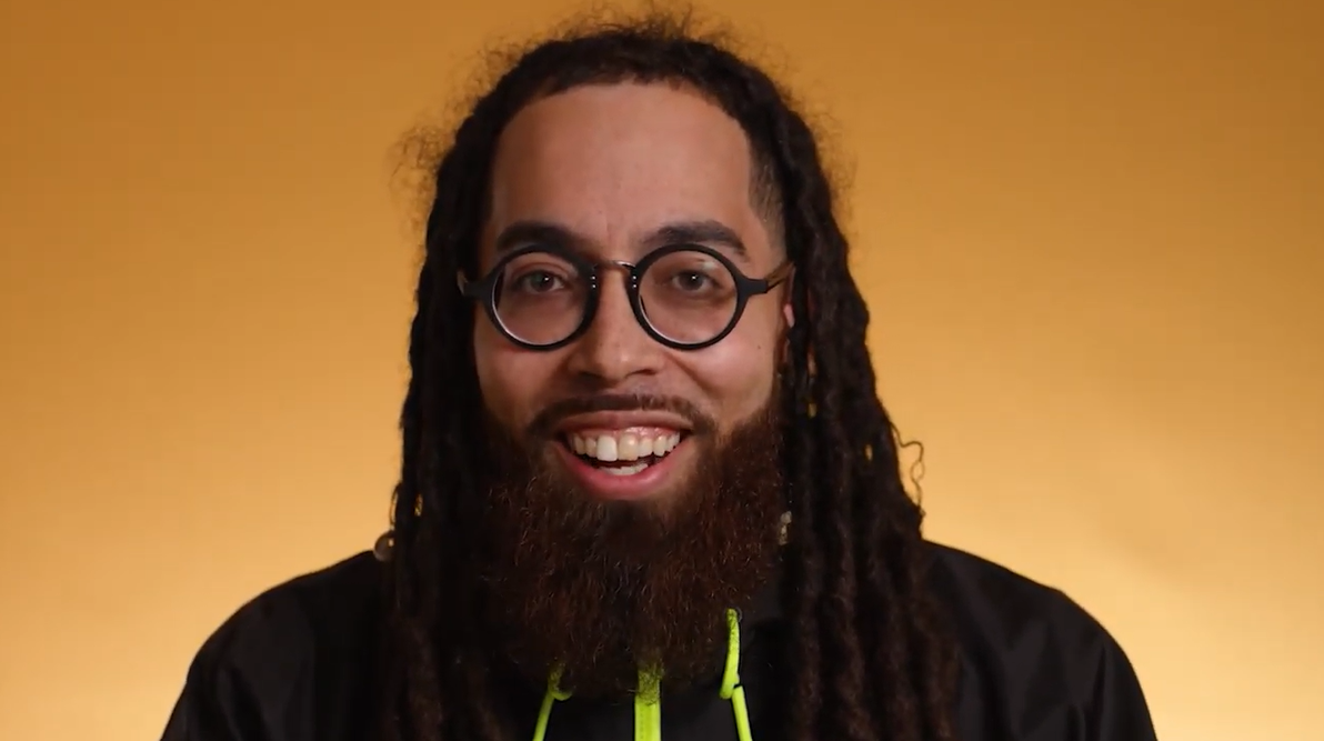 Back manwith facial hair, dreadlocks, and round glasses smiling mid-sentence at the camera.