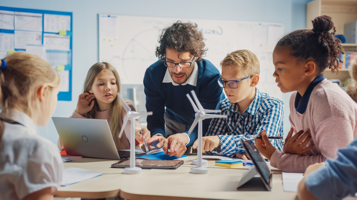 Young male teacher instructing a small diverse group of students using tablets and models of wind turbines.