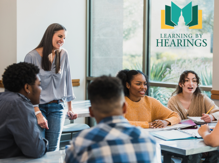 Teacher leaning on a desk chatting with high school students. the Learning by Hearings logo appears in the corner