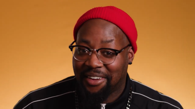 Black person with short facial hair and a red beanie talking to the camera